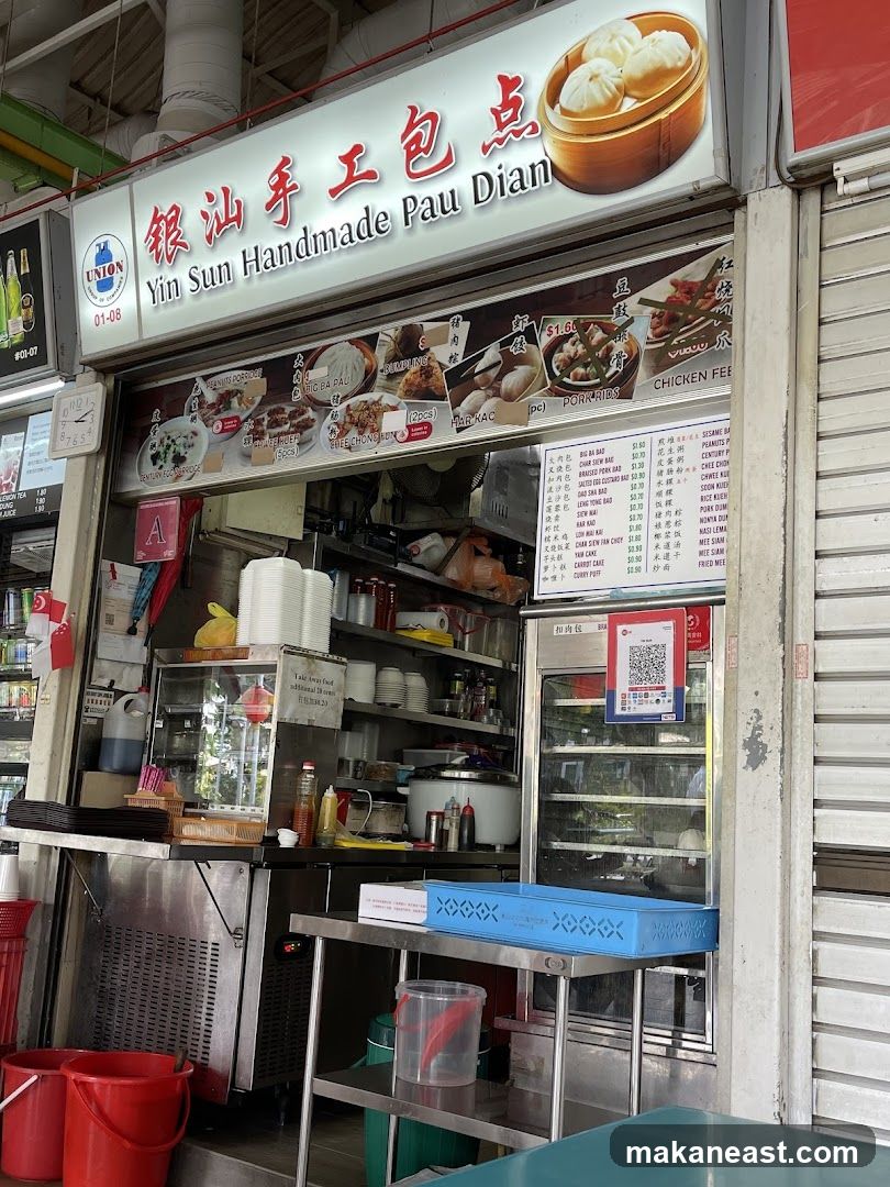 A food stall with a prominent sign displaying the business name and images of food items. The stall appears to be selling dim sum and other local dishes. There are menu boards and food displays visible.