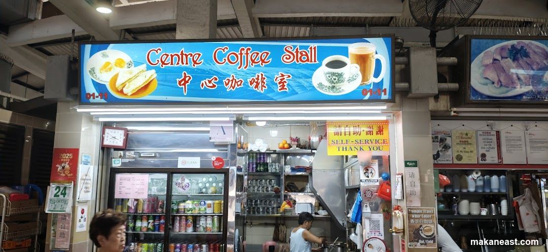 A coffee stall with a large illuminated sign displaying "Centre Coffee Stall" in English and Chinese, along with images of toast, eggs, coffee, and tea. Below the sign, there are refrigerators stocked with drinks, shelves with various items, and a person working behind the counter. A yellow sign reads "SELF-SERVICE THANK YOU". There are also several framed certificates and posters on the wall.