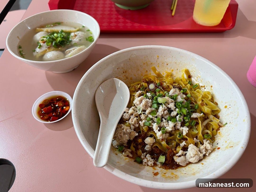 A close-up shot of a bowl of noodles topped with minced meat and chopped scallions, served with a side of fish ball soup and a small dish of chili. A spoon rests in the noodle bowl. In the background, a red tray holds chopsticks and a glass of yellow liquid.