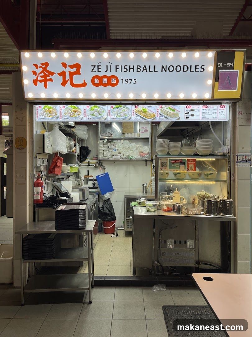 A brightly lit storefront for a fishball noodle shop. The sign above features the shop name in English and Chinese, along with images of their dishes and prices. The stall itself is visible behind the counter, showing kitchen equipment and food preparation areas.