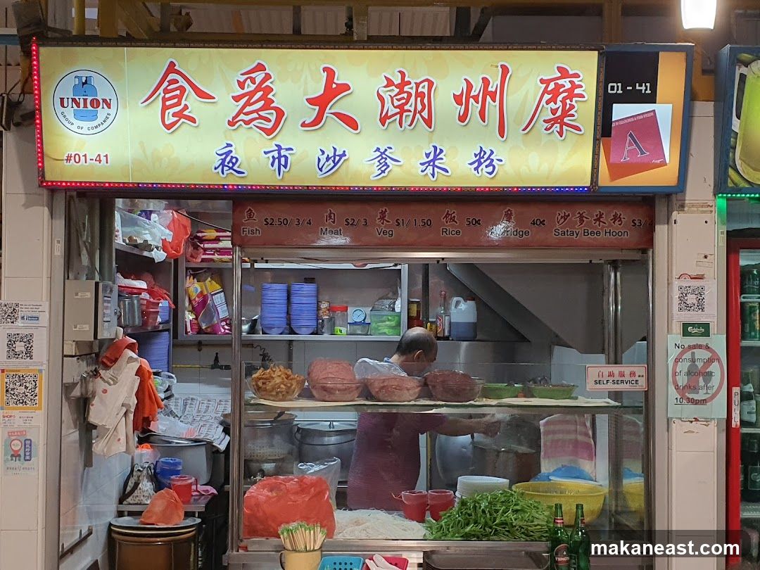 A food stall with a large illuminated sign displaying Chinese characters and the business name. Below the sign is a menu board with prices for various dishes. A person is visible working behind the counter. The stall appears to be in an outdoor or semi-outdoor setting, possibly a hawker center.