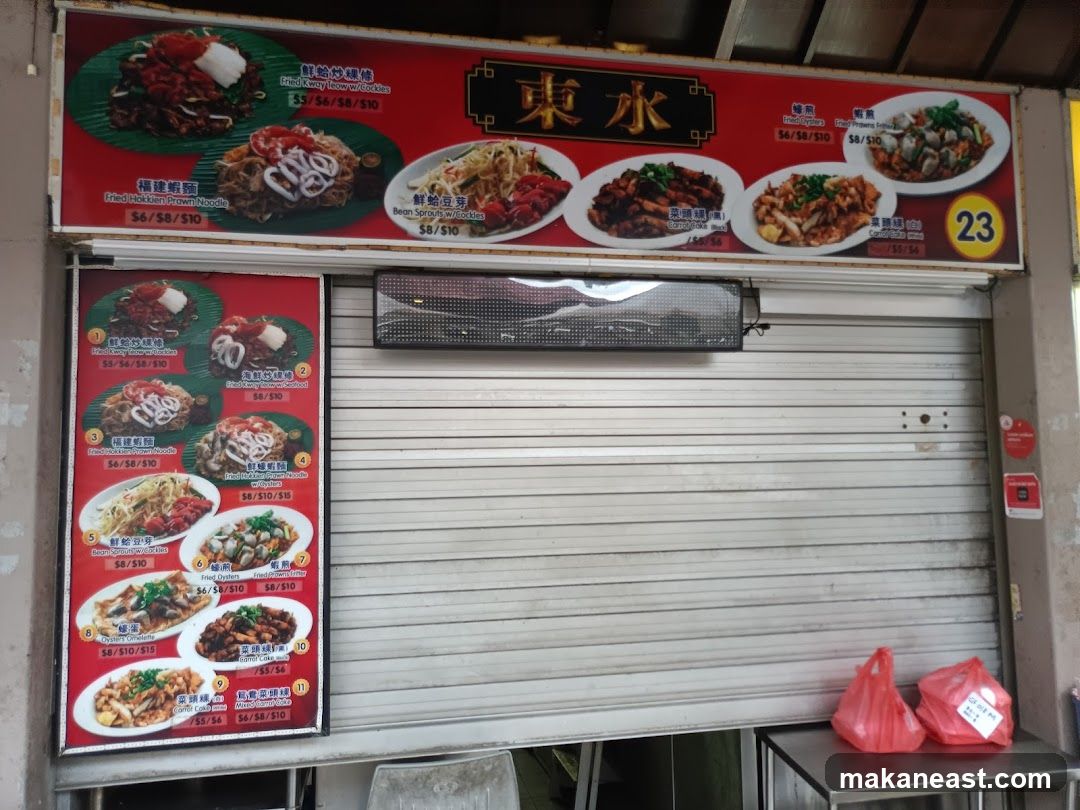 A food stall with a large red menu board displaying various noodle and seafood dishes. The stall has a metal roller shutter that is partially down. Two red plastic bags are on a metal table in front of the stall.