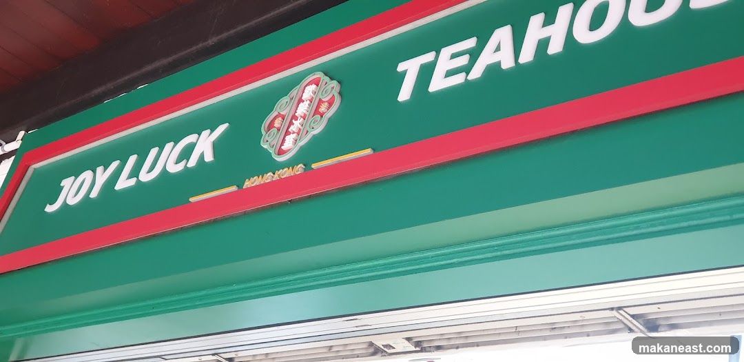 A close-up, angled shot of a green storefront sign with red trim. White 3D lettering spells out "JOY LUCK" on the left and "TEAHOUSE" on the right. In the center, a circular emblem with intricate designs and Chinese characters is visible, with the word "HONGKONG" below it.