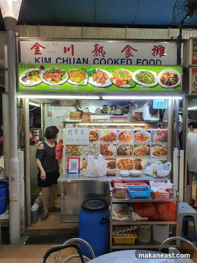 A street food stall with a large sign displaying the name "KIM CHUAN COOKED FOOD" in English and Chinese. Below the name, there are rows of pictures showcasing various dishes offered. A woman is standing inside the stall, and a QR code is visible on the counter. Shelving units with food containers and bags are also present.