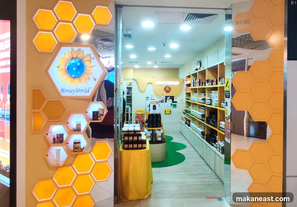 The exterior of a HoneyWorld store, featuring a prominent hexagonal display with a sunflower logo and the text 'HoneyWorld'. The display showcases various honey products in jars and bottles. Inside, shelves are stocked with more products, and a yellow tablecloth covers a display stand. The store's interior is brightly lit.
