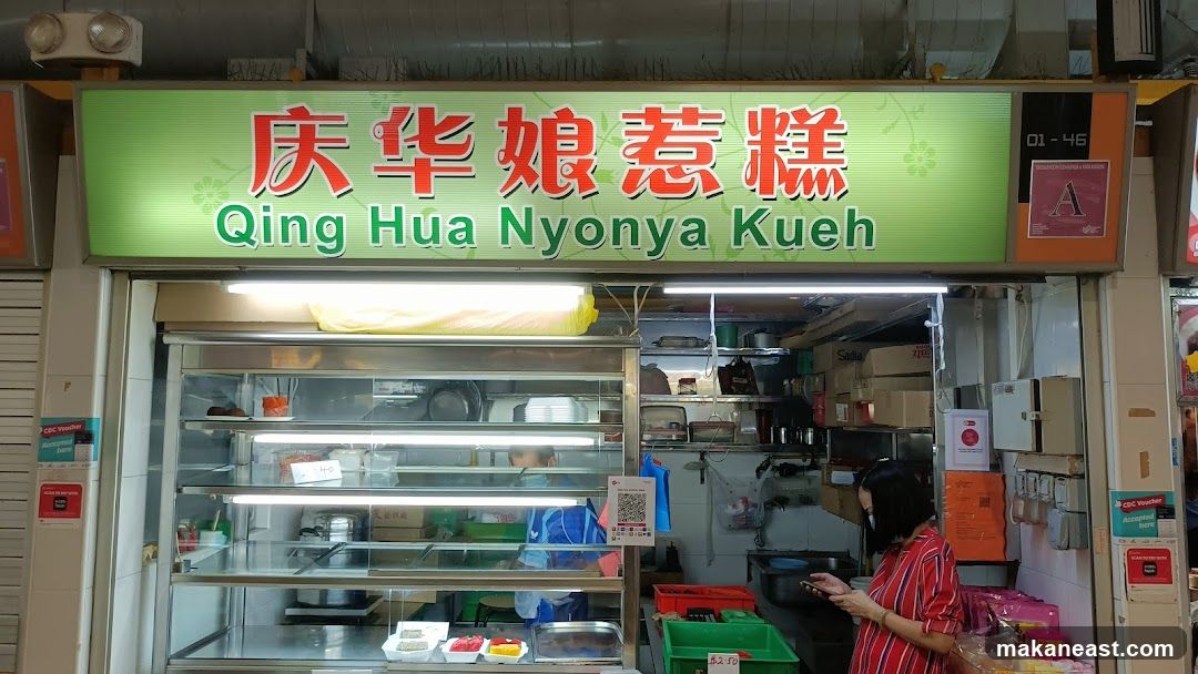A storefront with a bright green sign displaying Chinese characters and the English name "Qing Hua Nyonya Kueh". Below the sign is a display case filled with various colorful kueh (cakes/desserts). A woman in a red and white striped shirt is standing to the right, looking at her phone. The interior of the shop shows shelves stocked with boxes and containers.