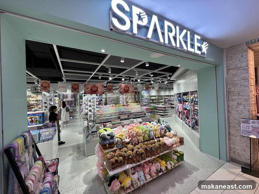 The exterior of a brightly lit store with a large illuminated sign that reads "SPARKLE" with a star graphic. The store entrance is wide, revealing shelves stocked with various items including plush toys, stationery, and accessories. Several sale signs are visible inside the store.