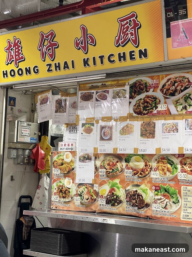 A food stall with a bright yellow sign displaying the name "HOONG ZHAI KITCHEN" in both English and Chinese. Below the sign, a menu board showcases various dishes with pictures and prices. The stall appears to be a self-service counter.