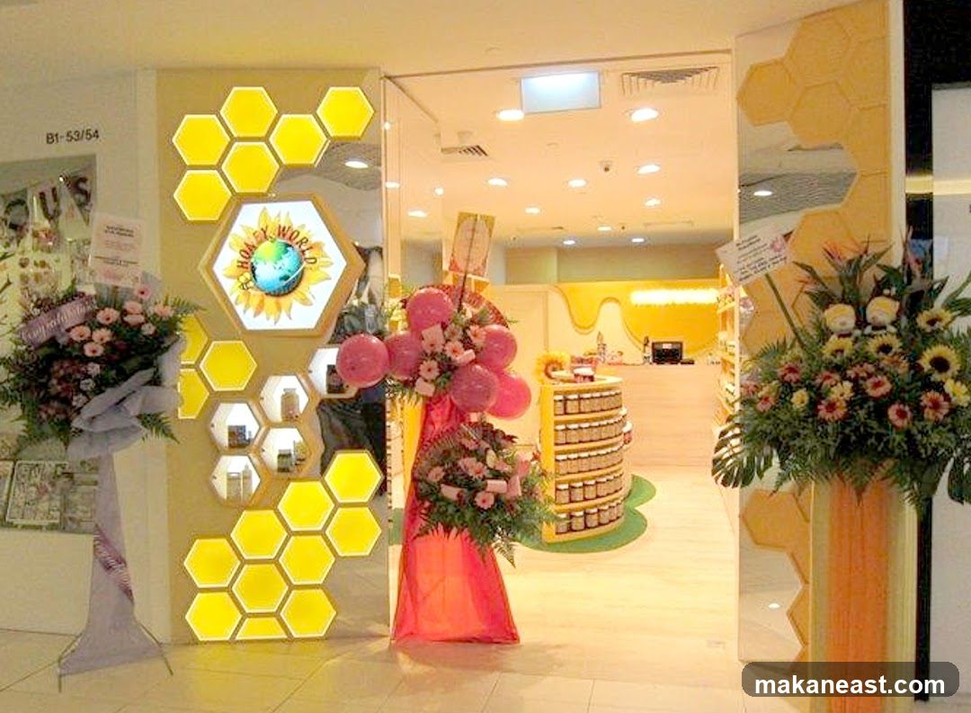 The entrance to a store called Honey World, decorated with honeycomb motifs and illuminated hexagonal panels. Floral arrangements and balloons are placed near the entrance, suggesting a special occasion. Inside, shelves stocked with jars are visible, along with a counter.