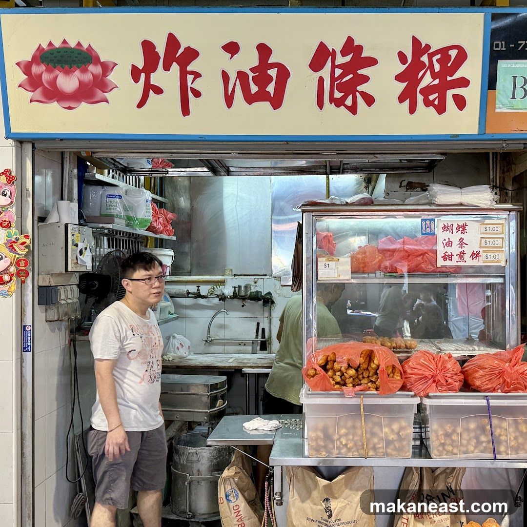 A man stands in front of a food stall with a large sign in Chinese characters and a lotus flower illustration. The stall displays fried dough fritters in plastic bags and containers. A glass display case shows more food items and a price list.