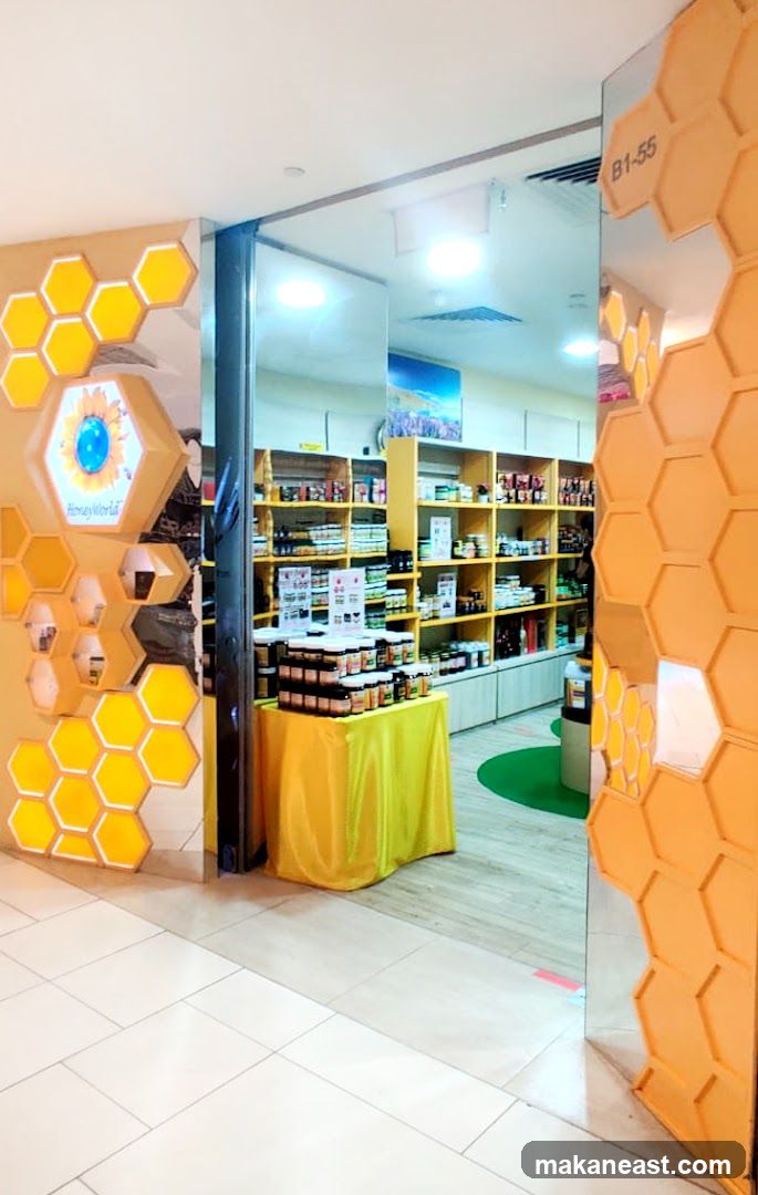 The exterior of a shop with a honeycomb-themed design. The entrance is open, revealing shelves stocked with jars of honey and other products. A yellow tablecloth drapes over a display table in front of the shelves. The shop's name, "Honey World," is visible on a hexagonal sign near the entrance.
