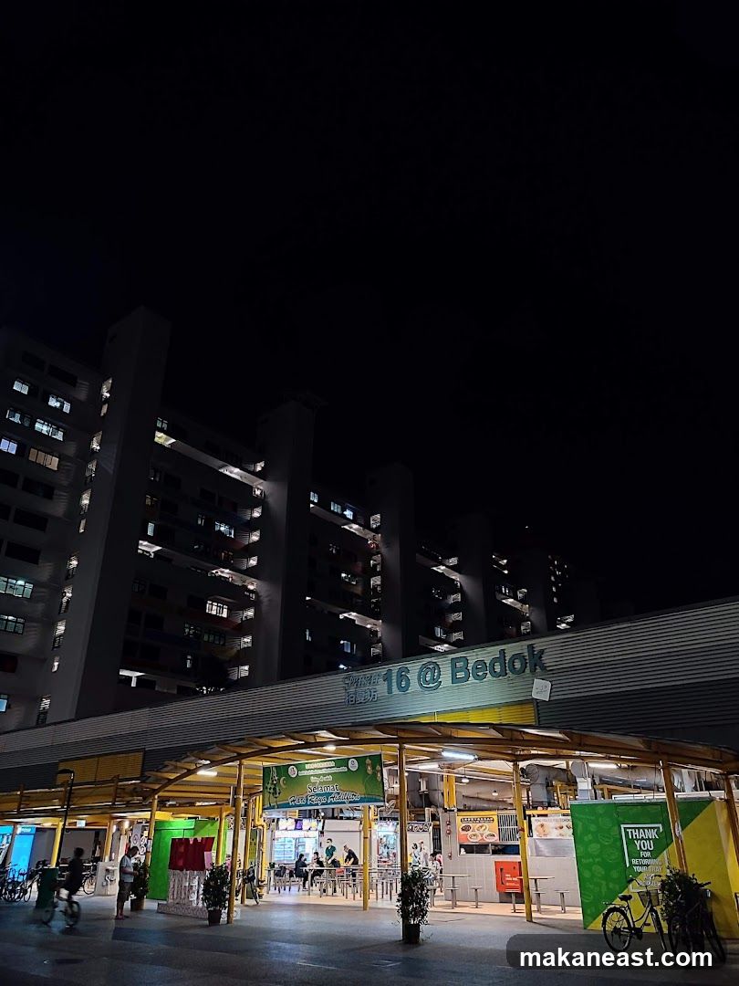 A nighttime view of a food court with a large apartment building in the background. The food court has a covered walkway with various stalls and seating areas. Signs indicate "16 @ Bedok" above the food court.