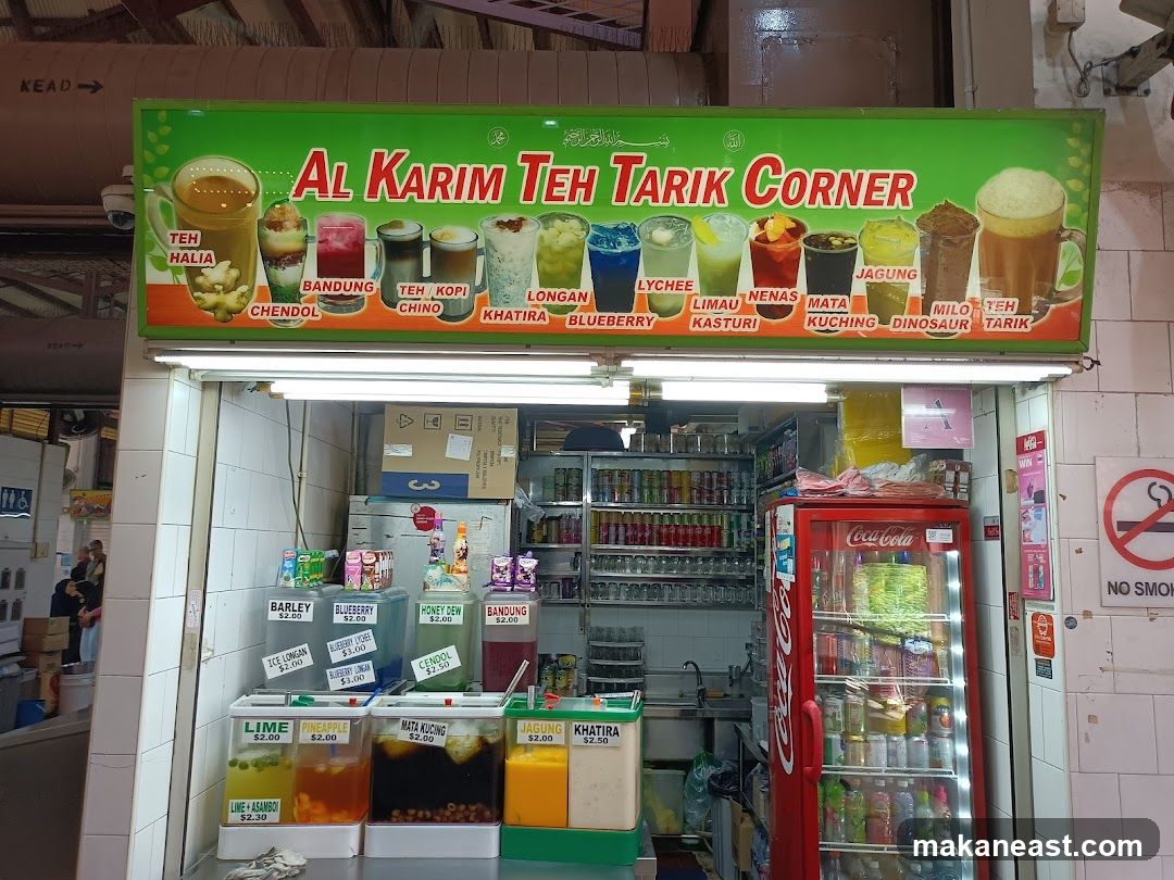 A storefront with a large green sign displaying various drinks and their names. Below the sign, there are several dispensers with drinks and prices, and a red Coca-Cola refrigerator. Shelves stocked with canned drinks are visible inside the shop.