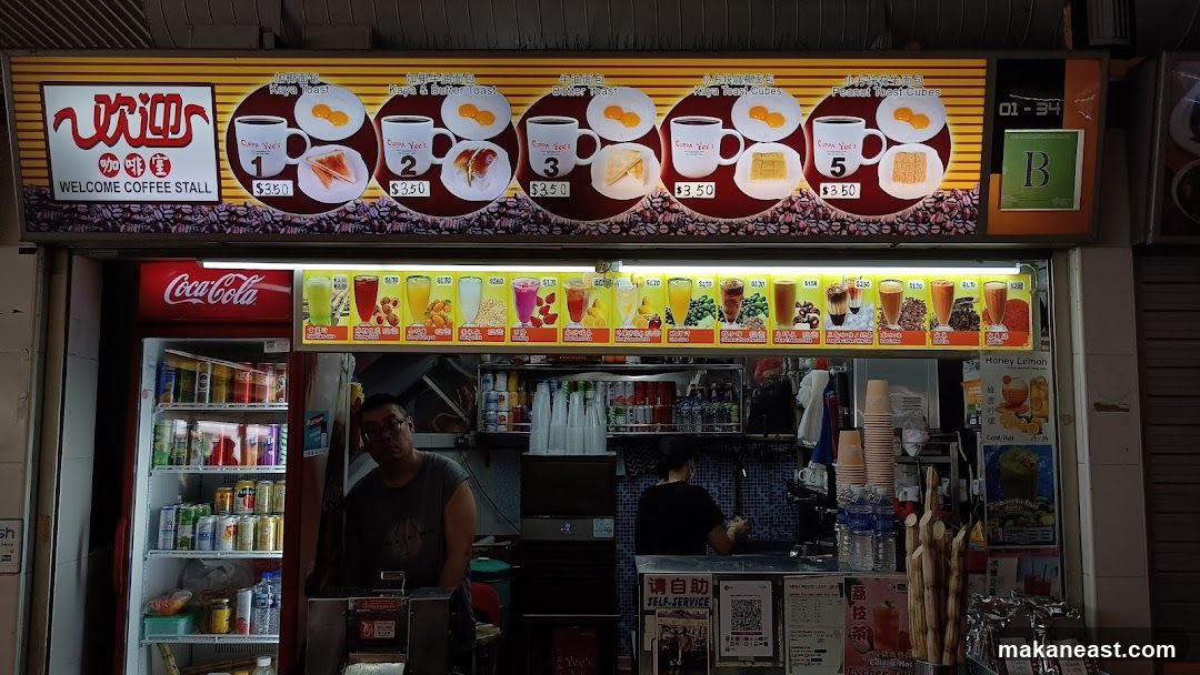 A coffee stall with a prominent sign displaying "WELCOME COFFEE STALL" in English and Chinese characters. The sign features images and prices of various breakfast items like Kaya Toast and Butter Toast, as well as drinks. Below the main sign is a Coca-Cola refrigerator stocked with beverages. The stall has a counter where staff are visible, and a menu board displaying different drinks with prices.