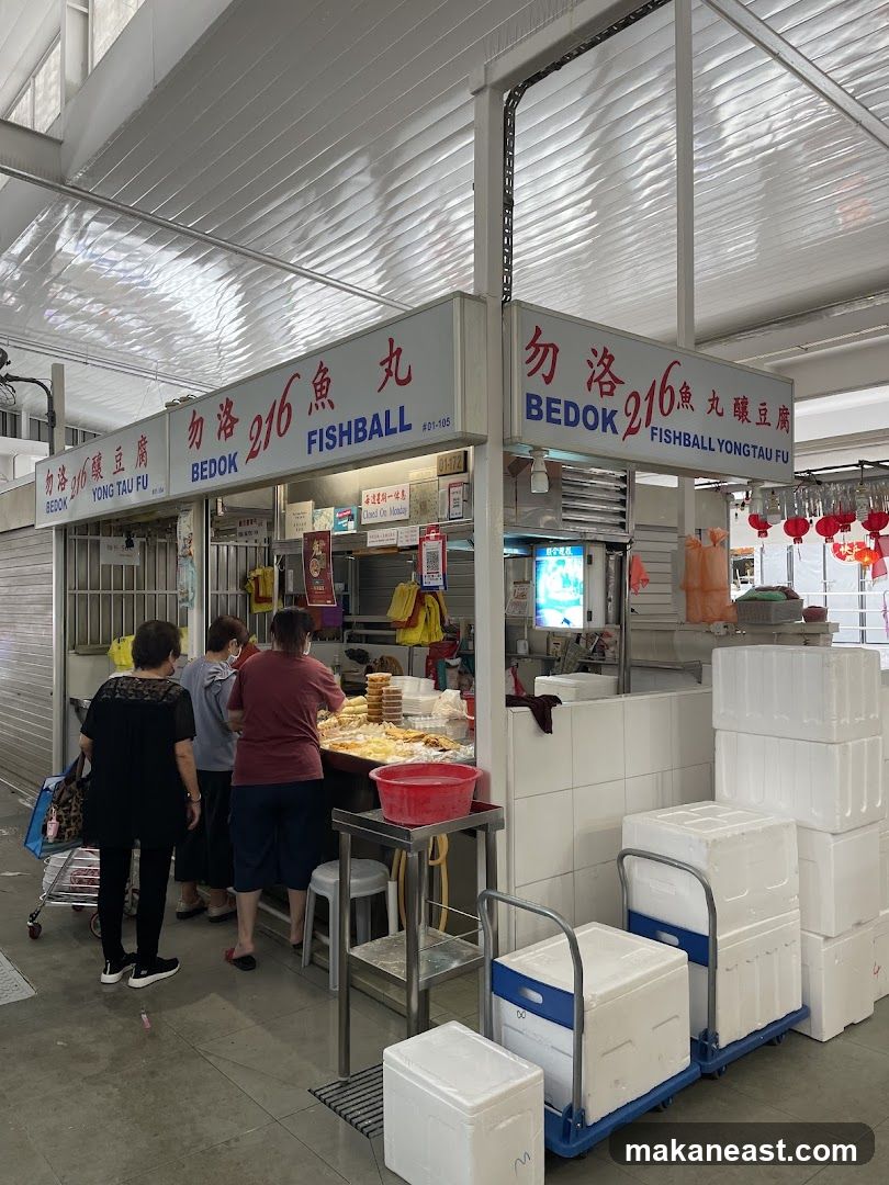 A food stall with prominent signage indicating "BEDOK 216 FISHBALL YONG TAU FU". Several customers are seen queuing and ordering food from the stall. The stall displays various food items and appears to be in a market or food court setting. There are also stacks of styrofoam boxes and trolleys outside the stall.