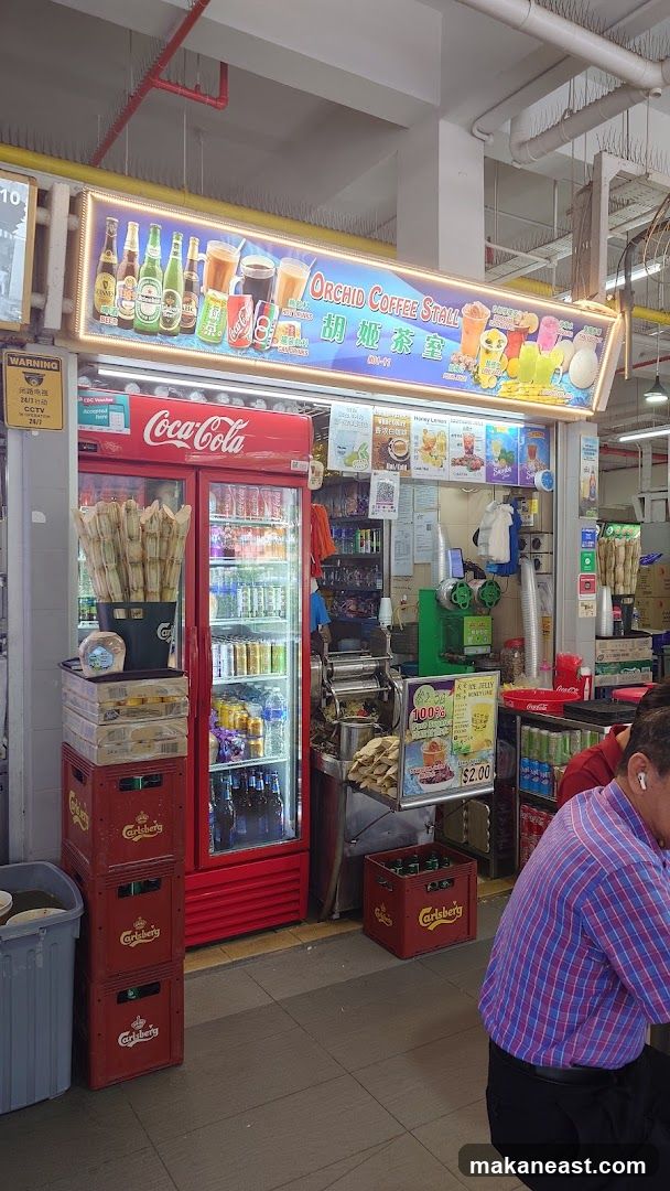 A coffee stall with a red Coca-Cola refrigerator displaying various beverages. Above the refrigerator is a brightly lit sign with the stall's name, "Orchid Coffee Stall," and images of drinks. Stacks of Carlsberg crates are visible next to the refrigerator. A sugarcane juicing machine is also present.