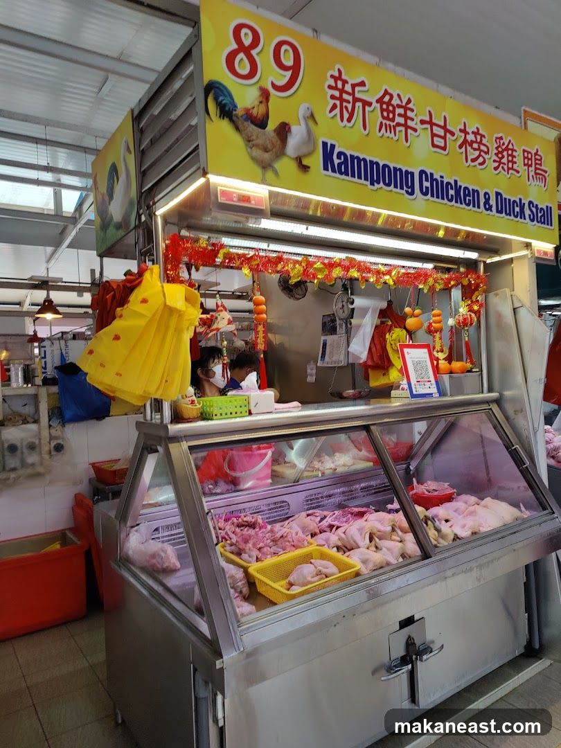 A stall with a large yellow sign displaying "89" and Chinese characters, along with the text "Kampong Chicken & Duck Stall". The sign features illustrations of chickens and ducks. Below the sign, a display case is filled with raw chicken and duck parts. Red and yellow decorations hang above the display. A person wearing a mask is visible behind the counter.