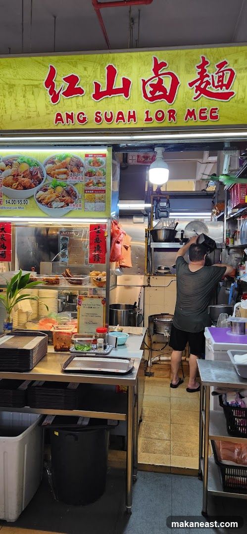 A food stall with a yellow sign featuring Chinese characters and the name "ANG SUAH LOR MEE" in English. Below the sign, there are pictures of food and prices. A person is working in the kitchen area behind the counter.