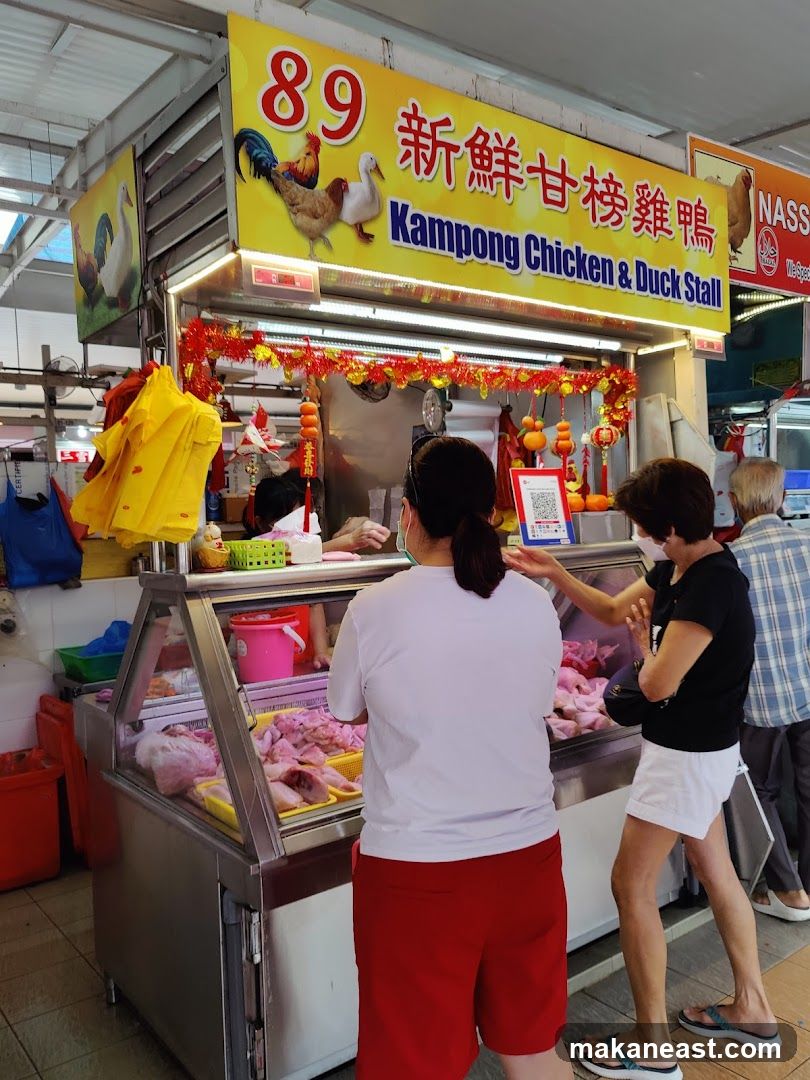 A stall with a bright yellow sign displaying "89" and Chinese characters, along with images of chickens and ducks. The sign reads "Kampong Chicken & Duck Stall". Below the sign, a display case contains raw chicken and duck parts. Two customers are visible, interacting with the stall owner. Red and gold festive decorations are hung above the display.