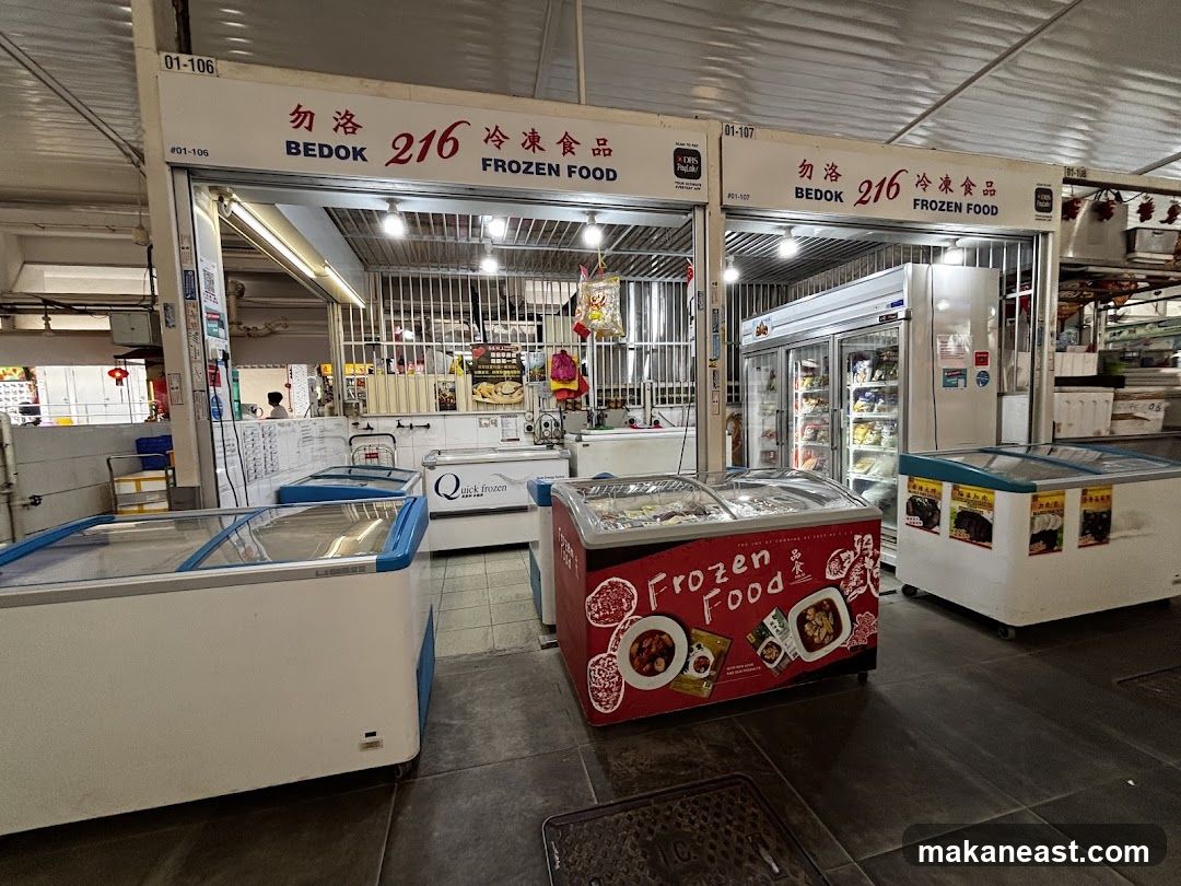 A storefront for a frozen food business named Bedok 216 Frozen Food. The facade features prominent signage with the business name in English and Chinese. Several chest freezers and upright refrigerators are visible outside the shop, displaying various frozen food items. The shop appears to be located in a market or food court setting.