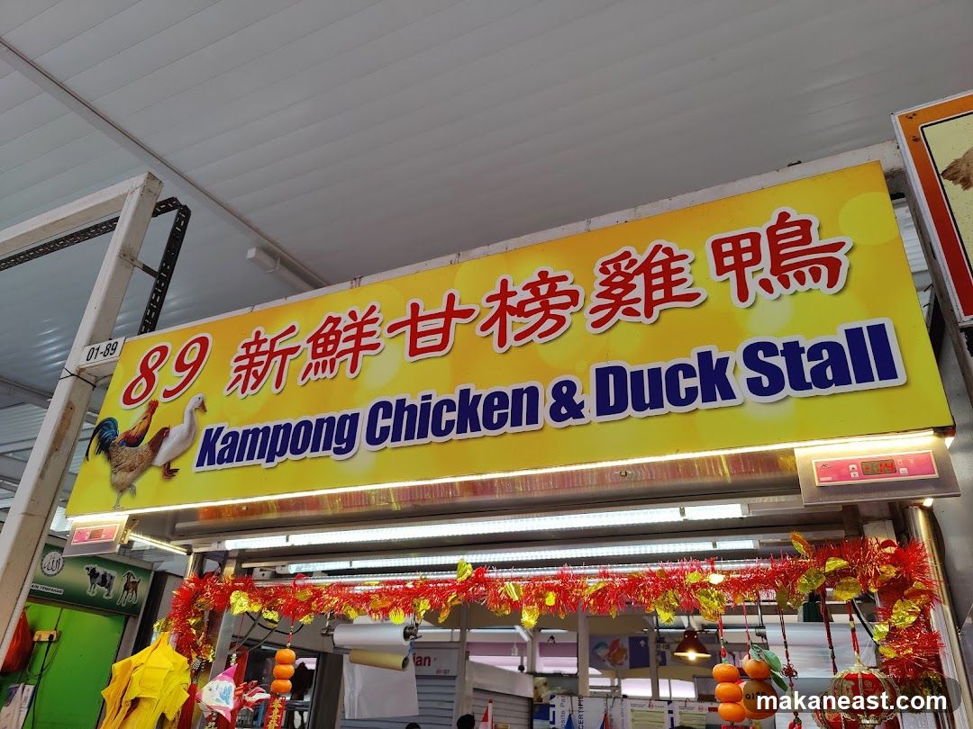 A bright yellow sign with red Chinese characters and blue English text reads "89 Kampong Chicken & Duck Stall". Images of a rooster and a duck are on the left side of the sign. Red and gold festive decorations hang below the sign.