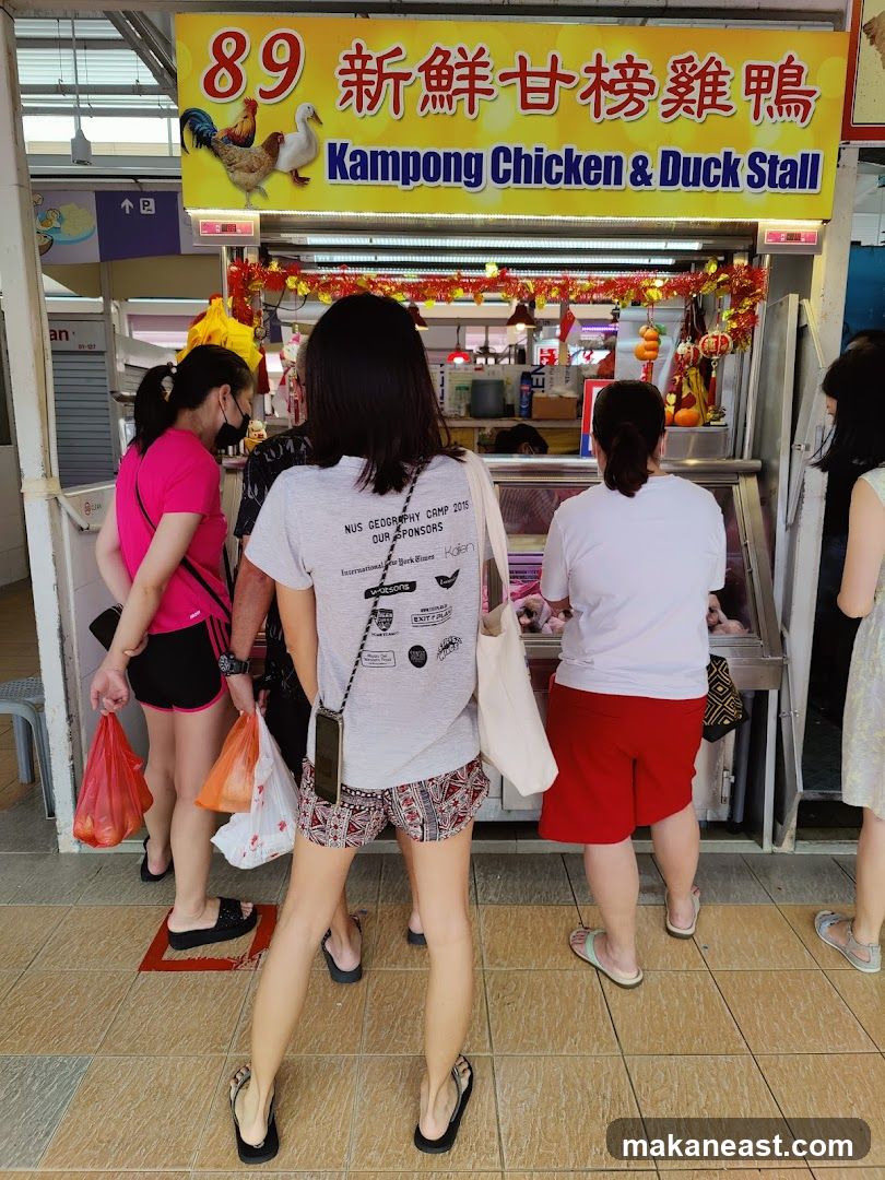 A stall with a yellow sign displaying "89 新鮮甘榜雞鴨 Kampong Chicken & Duck Stall" and images of a rooster and a duck. Several people are standing in front of the stall, some holding shopping bags. The stall appears to be in an open-air food court or market.