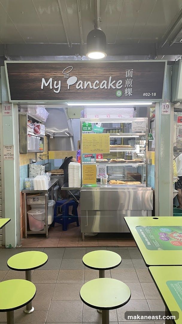 A food stall with a wooden sign that says "My Pancake" in English and Chinese characters. Below the sign is a display case with various food items. There are also tables and chairs in front of the stall.