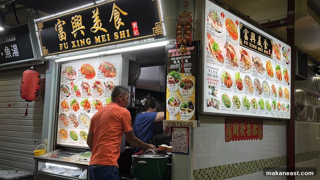 A food stall with a large illuminated menu board displaying various dishes. Two staff members are visible behind the counter. The stall has a prominent sign with Chinese characters and the name "FU XING MEI SHI".