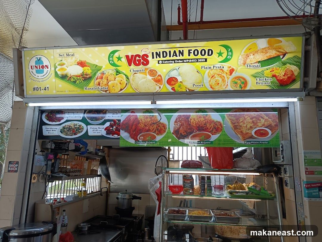 A food stall with a large yellow sign displaying various Indian dishes and the stall name "VGS INDIAN FOOD". Below the sign, a display showcases images of different food items with prices. The stall appears to be an outdoor or open-air establishment.