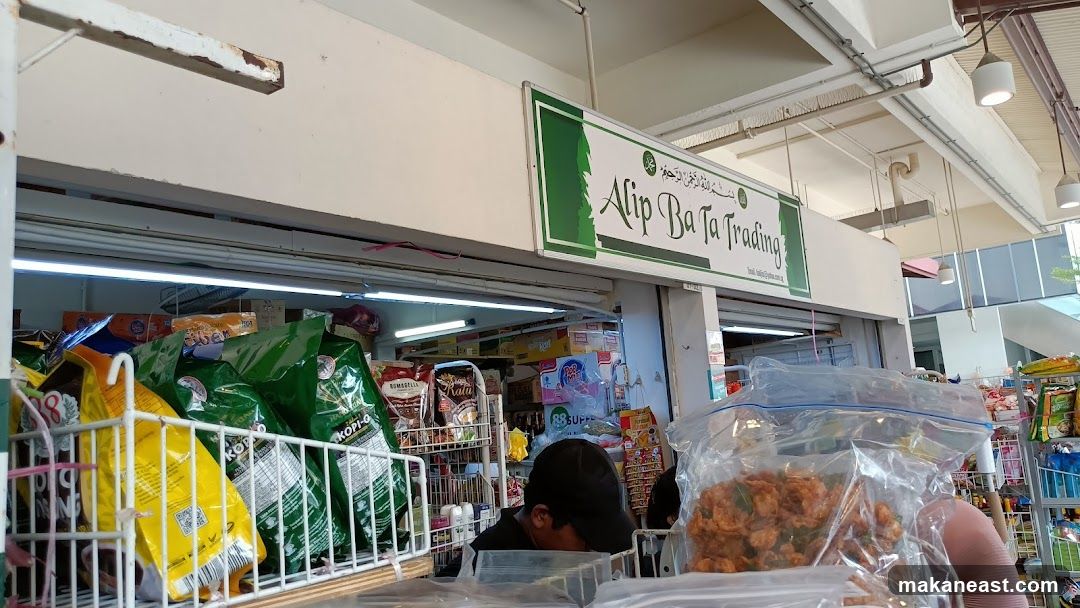 A view of the storefront of Alip Ba Ta Trading, a small grocery or convenience store. The store is filled with various packaged goods, including bags of coffee, snacks, and other food items. A sign above the entrance reads "Alip Ba Ta Trading" with green decorative elements. The interior is visible, showing shelves stocked with products and a person working behind the counter.