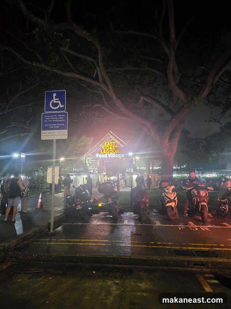A nighttime exterior view of the East Coast Lagoon Food Village. A sign indicating reserved parking for handicapped vehicles is visible on the left. Several motorcycles are parked in front of the food village, which is illuminated by bright lights. People are seen milling around the entrance.