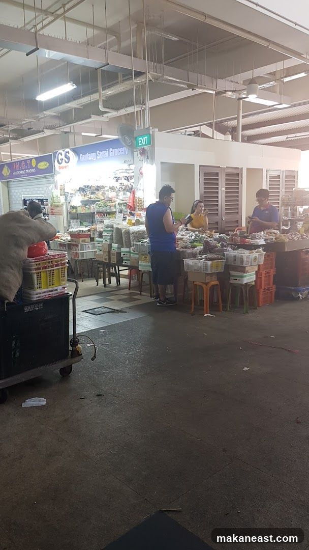 A bustling market scene with various stalls displaying a wide array of goods, including produce, dried goods, and other items. People are seen interacting with vendors and browsing the merchandise. An 'EXIT' sign is visible above one of the stalls.