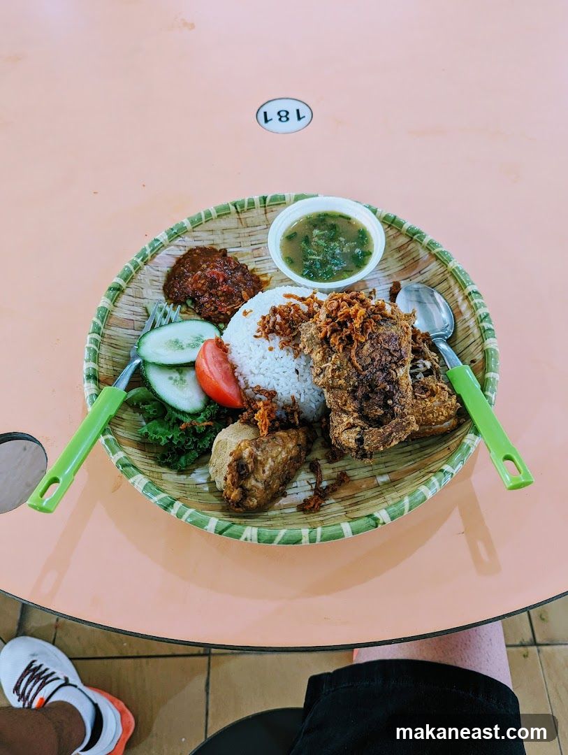 A close-up overhead shot of a meal served on a woven platter. The meal includes white rice, a piece of fried chicken, a small dish of chili paste, sliced cucumbers, a tomato slice, lettuce, a piece of fried tofu or tempeh, and a small bowl of soup or sauce. The platter is placed on a light orange table. A table number '181' is visible in the background. A person's legs and feet wearing sneakers are visible at the bottom of the frame.