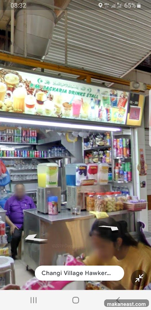 A drinks stall with a sign displaying various beverages and the stall name. Shelves behind the counter are stocked with canned drinks and bottled water. A man in a purple shirt is seated to the left, and a person is blurred in the foreground.