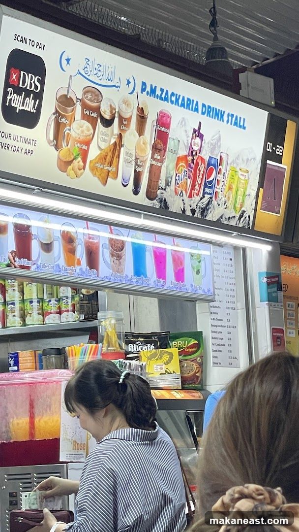 A drink stall with a large illuminated menu board displaying various drinks and some food items. Below the menu board, there are shelves stocked with canned drinks and other products. A woman is seen in the foreground, handling money.