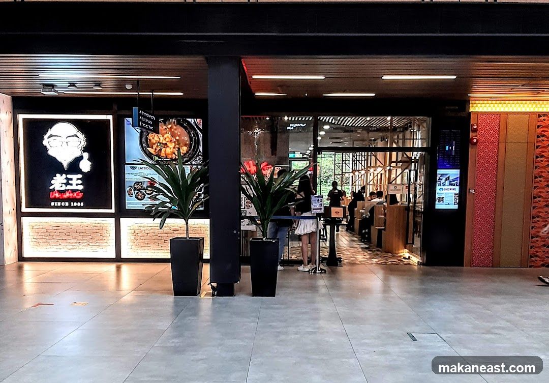 A restaurant storefront with a large illuminated sign featuring a cartoon character and Chinese characters, next to a display of food. Inside, diners are seated at tables.