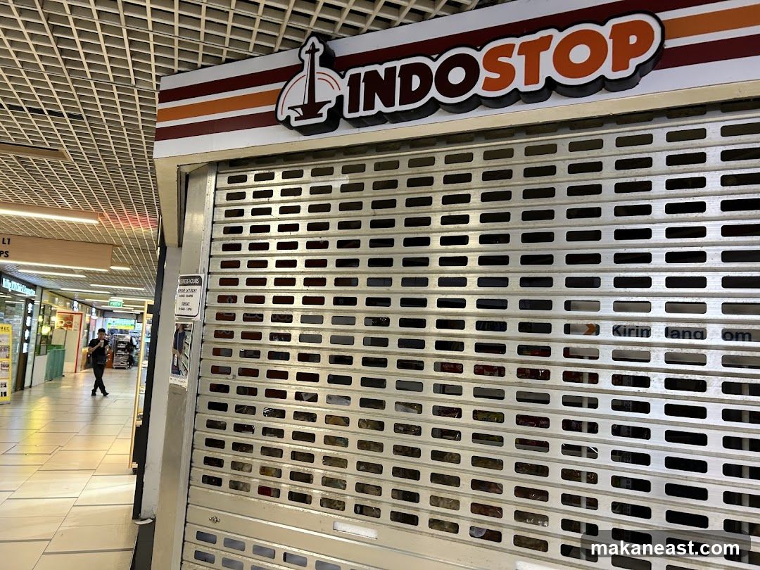 A closed storefront with a metal security shutter. The sign above reads "Indostop" with a graphic of a monument. A business hours sign is visible on the left side of the shutter. The interior of the shop is dimly lit and stocked with products. A person is walking in the hallway outside the shop.