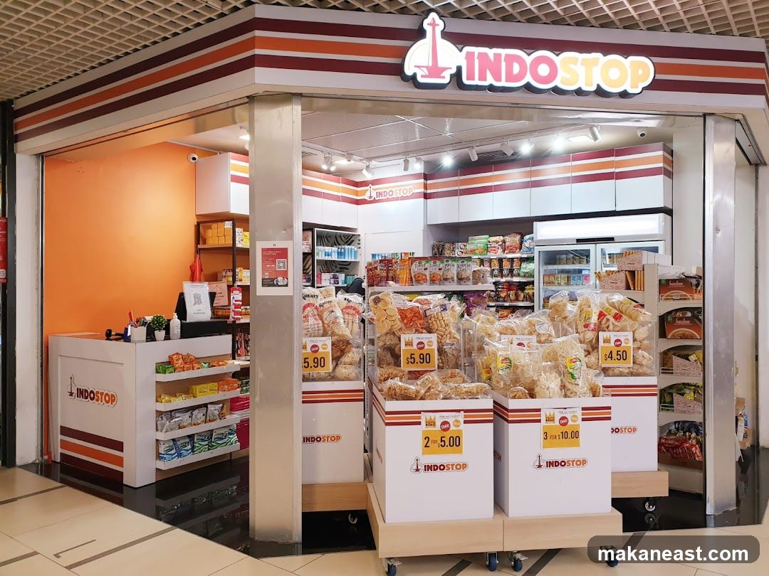 A brightly lit convenience store with an orange accent wall and a white counter. Shelves behind the counter are stocked with various products. In front of the store, several display boxes are filled with bagged snacks. The store's name, "INDOSTOP," is prominently displayed on a sign above the entrance and on the counter and display boxes. Prices are visible on some of the snack displays.