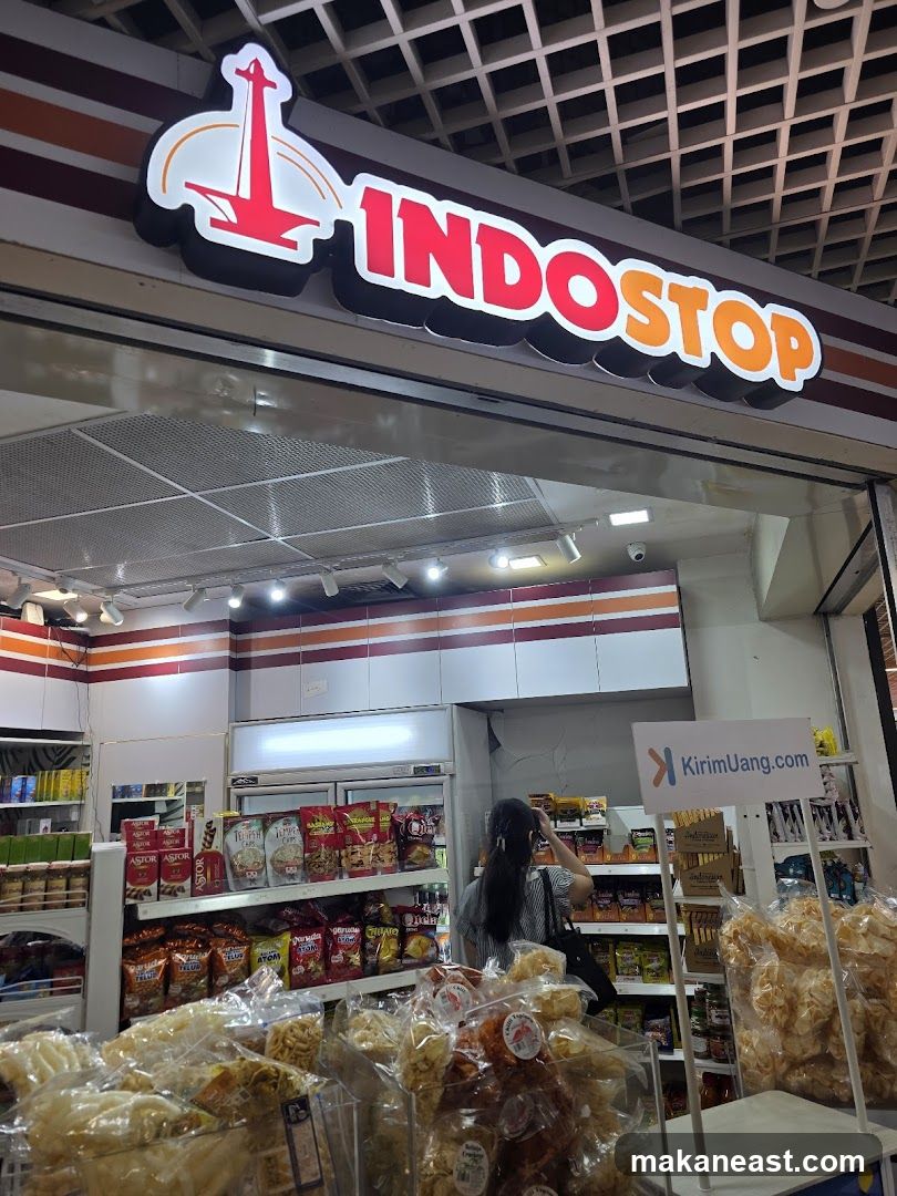 The image shows the exterior of a store called Indostop. The sign is illuminated with red and orange letters. Inside the store, shelves are stocked with various packaged goods, including snacks and drinks. A woman is browsing the products. In the foreground, there are large displays of bagged snacks. A sign for "KirimUang.com" is visible.