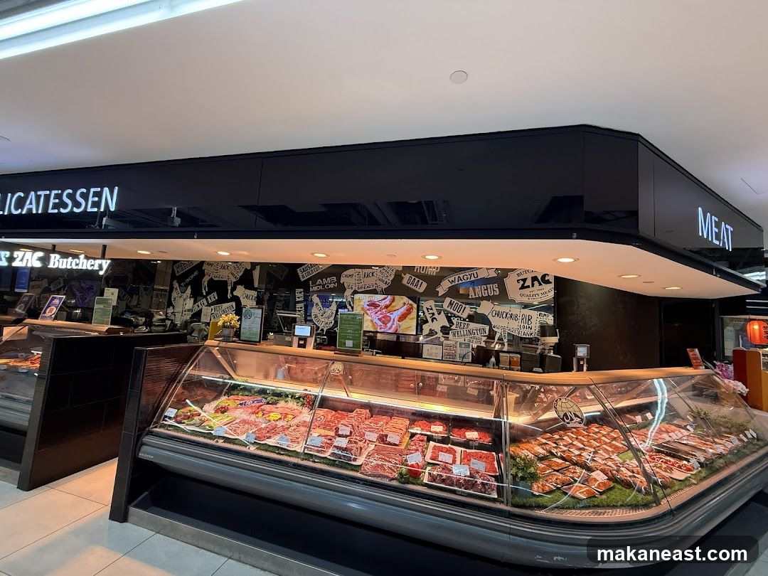 A brightly lit delicatessen and meat shop with a large display case filled with various cuts of meat. The shop has signs that read "DELICATESSEN" and "MEAT". The interior features a black wall with white text and graphics related to meat cuts and breeds.