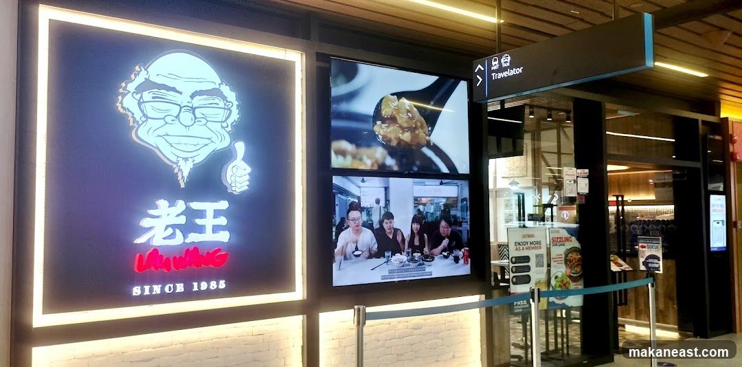 A brightly lit storefront features a large neon sign with a cartoon caricature of an elderly man and Chinese characters, along with the text "SINCE 1985". To the right of the sign are two digital screens displaying food and people dining. A directional sign for "Travelator" is visible above the entrance. Several promotional posters are displayed near the entrance, including one advertising "Sizzling" dishes and another encouraging membership.