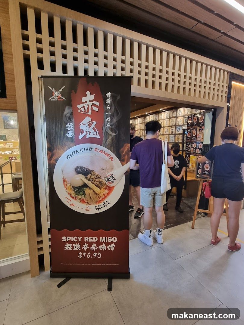 A tall banner displays a bowl of ramen and text in Japanese and English. The text includes "Shinsho Ramen" and "Spicy Red Miso $16.90". In the background, people are standing near the entrance of a restaurant with a wall covered in what appear to be small wooden boxes or signs.