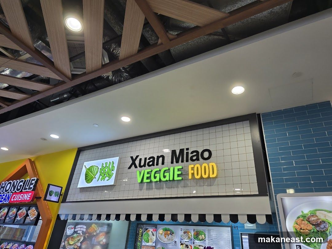 A sign for "Xuan Miao Veggie Food" is displayed on a tiled wall. To the left, a sign for "Hong Le Korean Cuisine" is visible. Below the signs, a menu board displays various food items. The ceiling features exposed wooden beams and recessed lighting.
