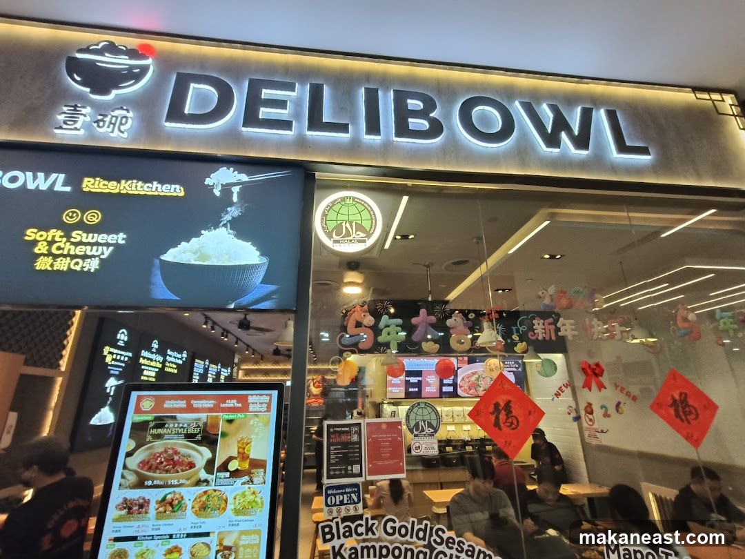 The storefront of DELIBOWL Rice Kitchen features a large illuminated sign with the restaurant's name. To the left, a digital display shows an image of rice with the text "Soft, Sweet & Chewy". A Halal certification logo is visible on the right. Inside, customers are seated at tables, and decorations for the New Year are displayed.