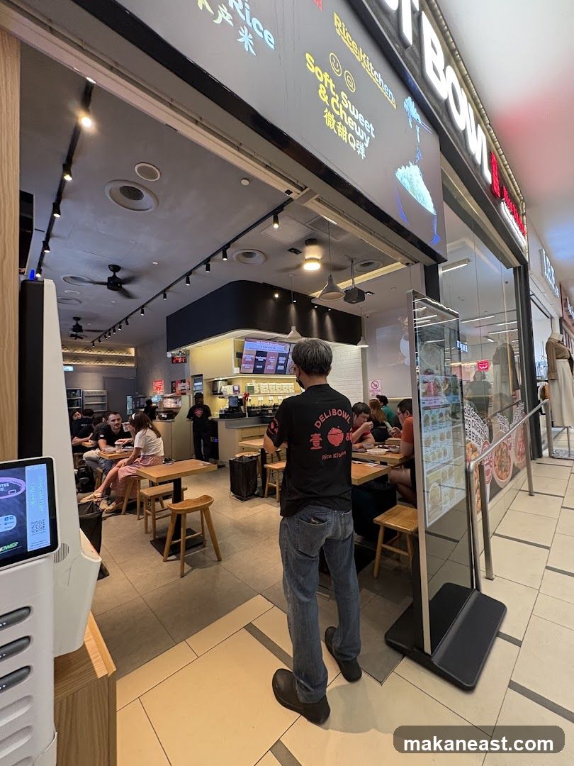 A view of the exterior of a restaurant with a prominent sign that reads "DeliBowl Rice Kitchen". The restaurant has an open-concept seating area visible through large glass windows. Inside, customers are seated at tables, and staff are visible behind the counter. A digital menu board is also visible.