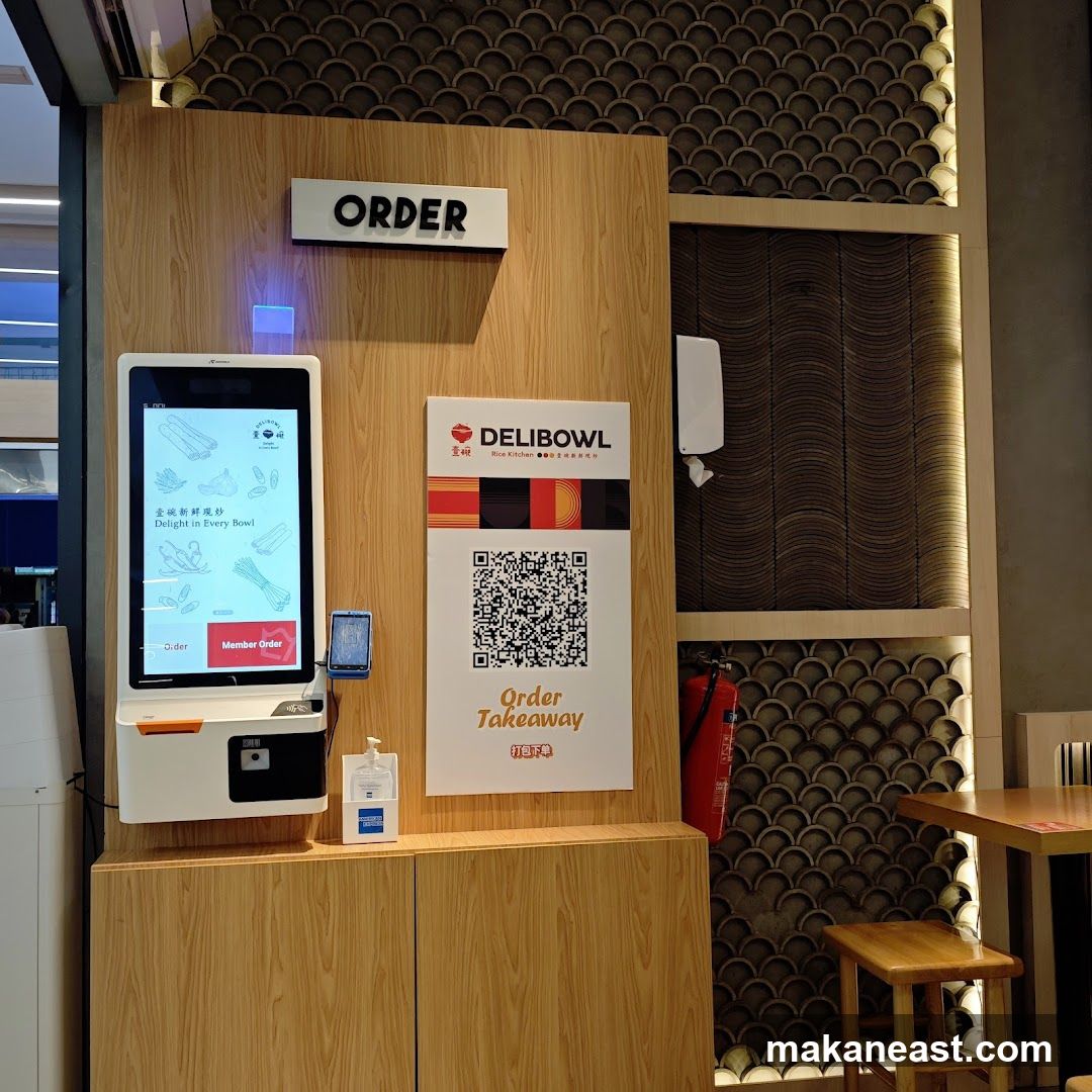 A wooden counter with a digital ordering kiosk and a sign for DELIBOWL. The kiosk displays images of ingredients and text in Chinese and English. A QR code for ordering takeaway is also visible. Hand sanitizer is mounted on the wall. A fire extinguisher is to the right.